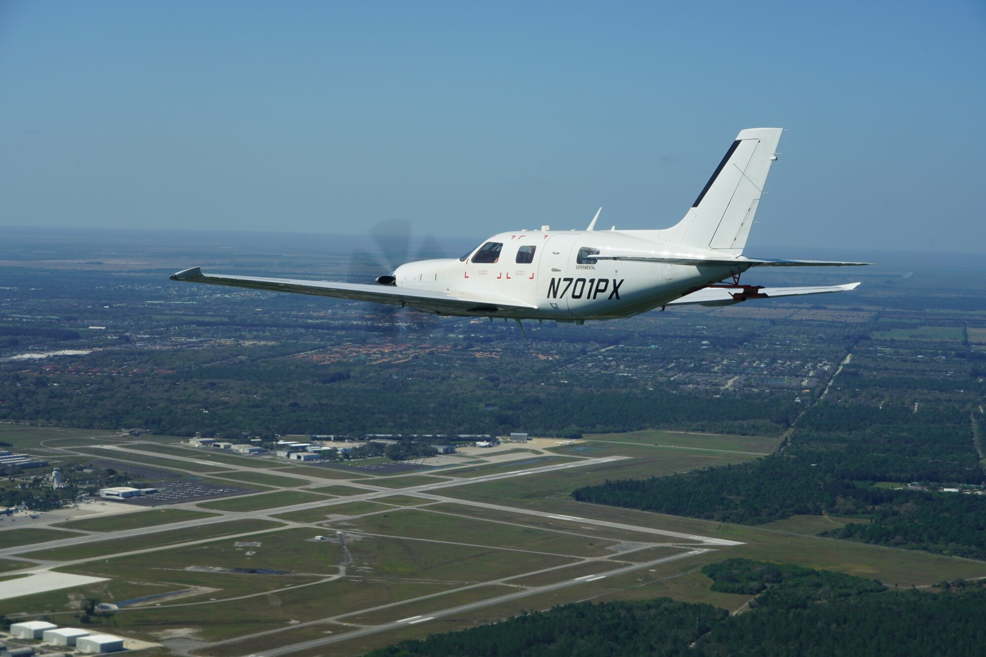 A Piper M700 aircraft flying over an airport runway, with a clear blue sky in the background.