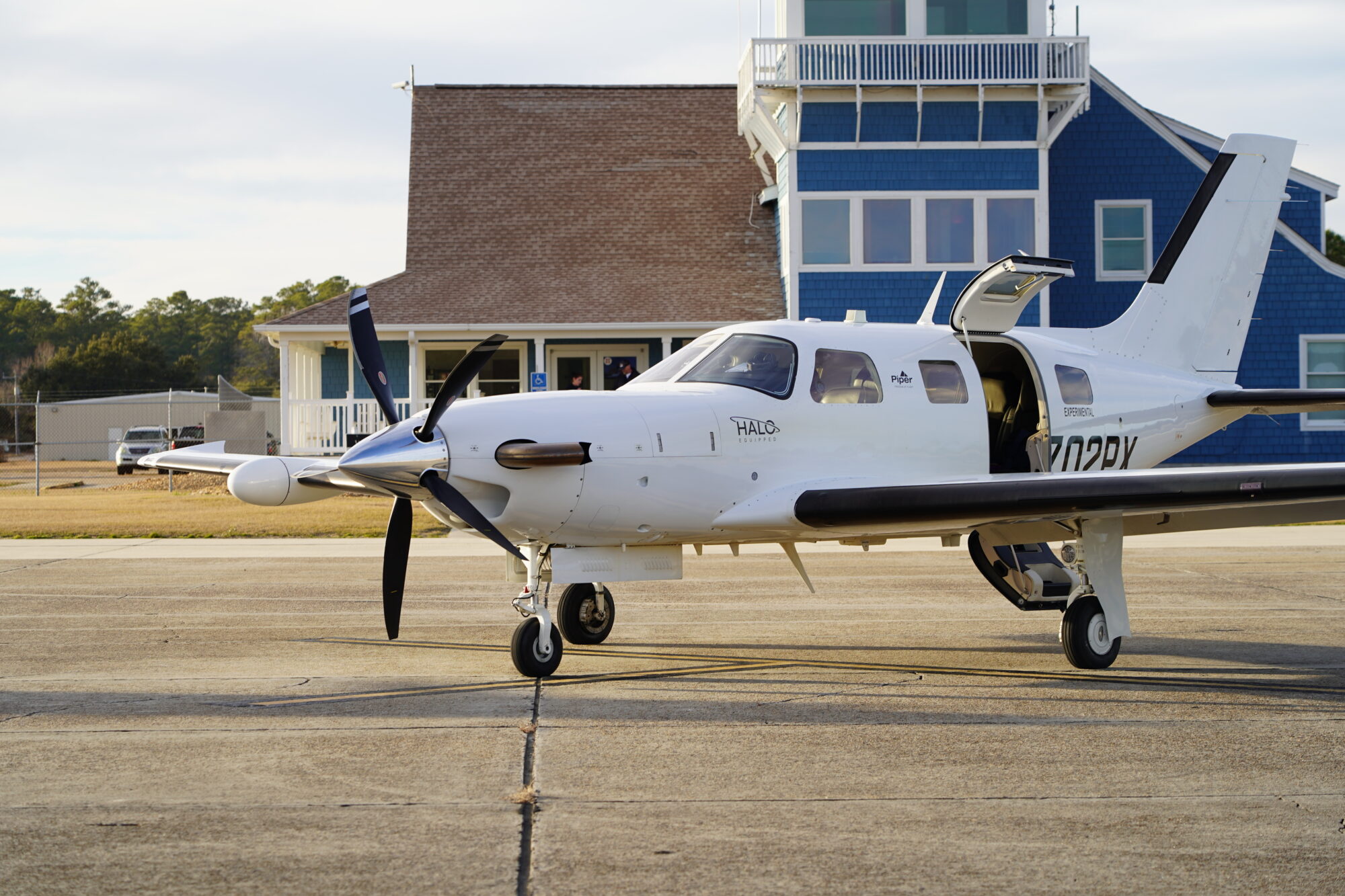 A Piper M700 parked on the tarmac with its door open, set against a building with a pitched roof.