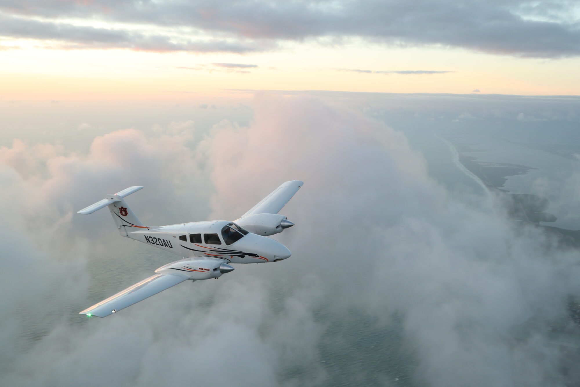 A Piper Seminole twin-engine aircraft in flight above a cloud-covered coastline during sunset, with the shoreline visible through breaks in the clouds.