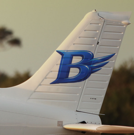 Close-up of a white airplane tail at sunset featuring Blitz Aviation’s blue ‘B’ logo with stylized wings, with blurred trees in the background.