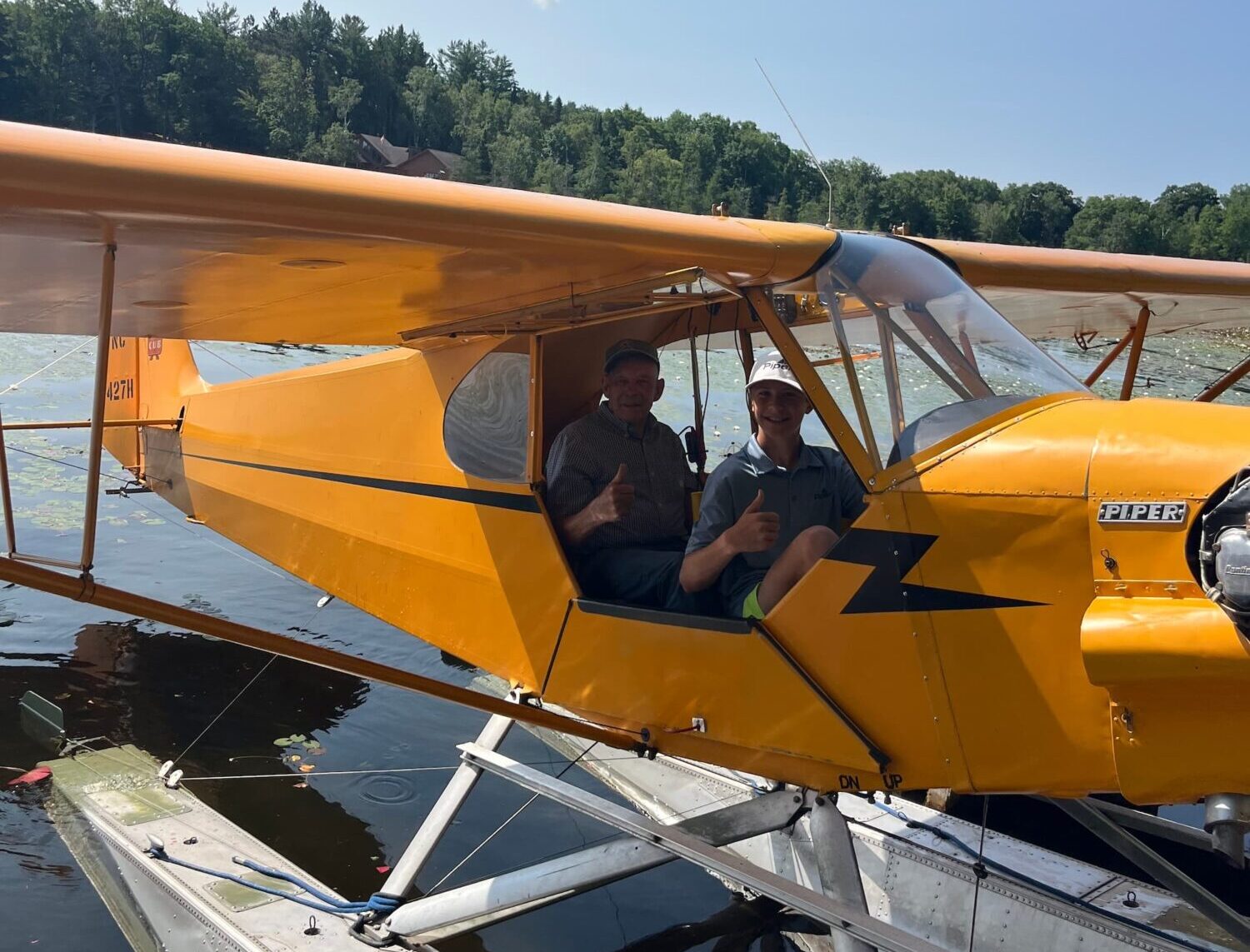 Two people sitting in a yellow Piper J-3 Cub NC7427H give a thumbs up from the open cockpit while the airplane sits on floats on a calm lake.