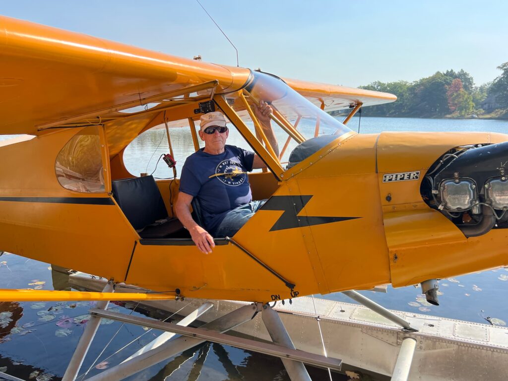 A man sits in the open cockpit of yellow Piper J-3 Cub NC7427H on floats, moored on a calm lake among lily pads.
