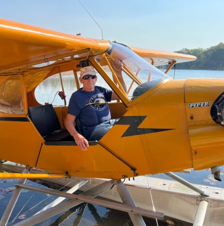 A man sits in the open cockpit of yellow Piper J-3 Cub NC7427H on floats, moored on a calm lake among lily pads.