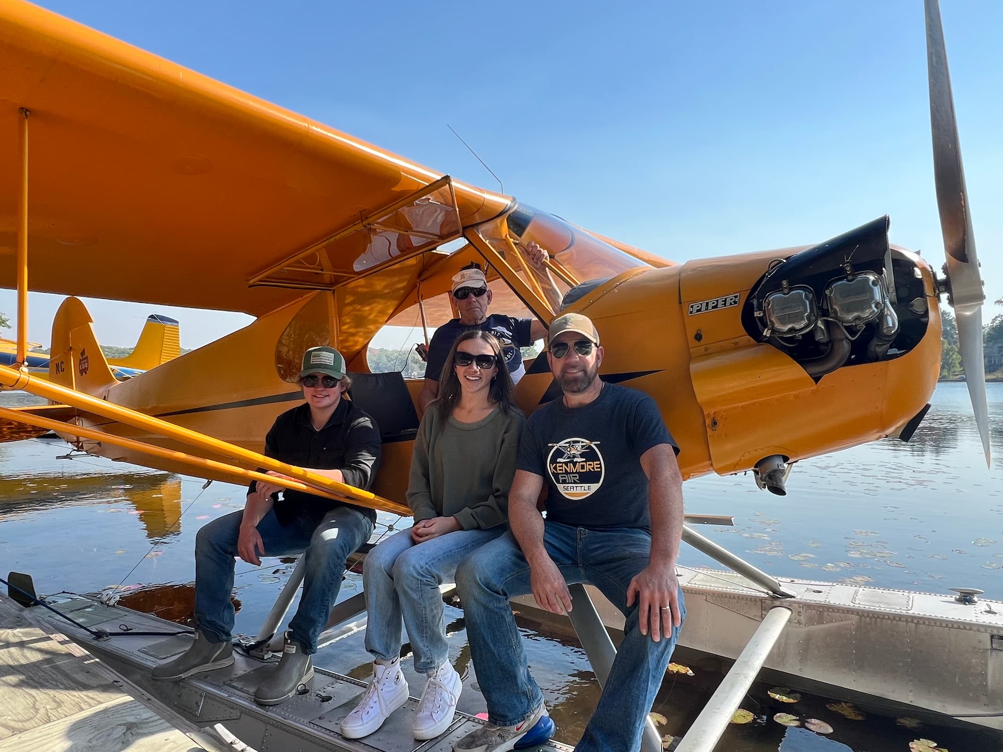 NC7427H: A Piper J-3 Cub's Journey 7 Three people sit side by side on the float of yellow Piper J-3 Cub NC7427H while a fourth stands in the open cockpit doorway above them.