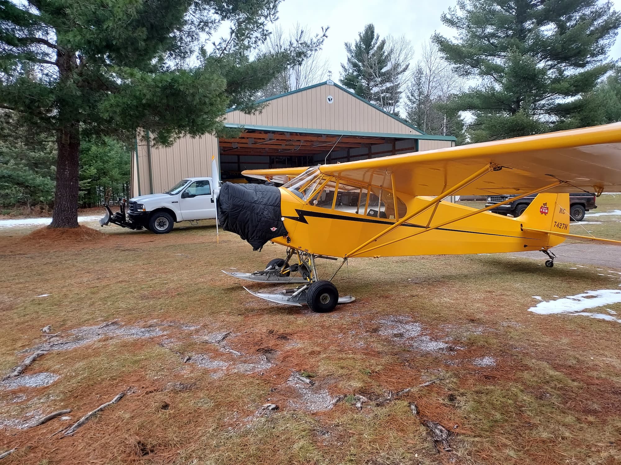 NC7427H: A Piper J-3 Cub's Journey 4 Piper J-3 Cub NC7427H on skis, parked on frosty ground outside a tan hangar, an insulated cover over its engine.