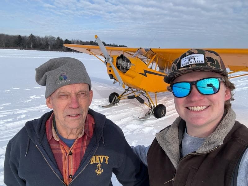 NC7427H: A Piper J-3 Cub's Journey 6 Don in a veteran's beanie and a younger man in a ball cap and sunglasses stand together on snow, with a yellow Piper J-3 Cub on skis parked behind them.